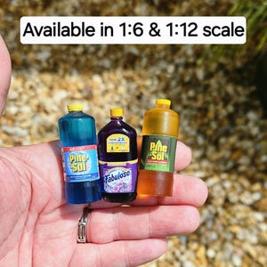 May include: Miniature cleaning product bottles, including Pine-Sol and Fabuloso, are displayed. The bottles are in various colors, with labels and yellow caps. The text "Available in 1:6 & 1:12 scale" is visible. These are likely for dollhouses or miniature collections.