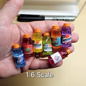 Pu&ograve; includere: Miniature bottiglie di Gatorade in vari colori, tra cui blu, rosso, giallo, verde, arancione e viola, con tappi arancioni. Le bottiglie sono tenute in una mano e il testo "1:6 Scale" &egrave; visibile.