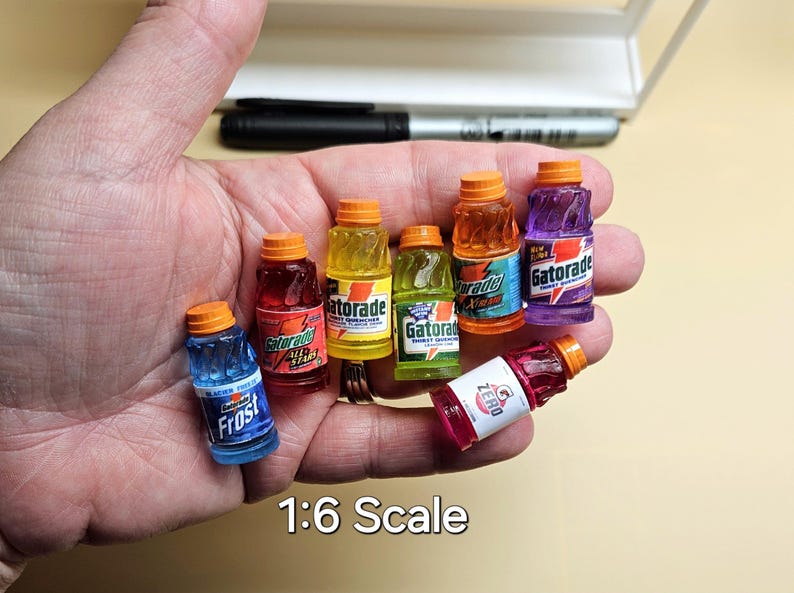Pu&ograve; includere: Miniature bottiglie di Gatorade in vari colori, tra cui blu, rosso, giallo, verde, arancione e viola, con tappi arancioni. Le bottiglie sono tenute in una mano e il testo "1:6 Scale" &egrave; visibile.