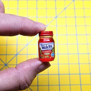 May include: A miniature glass jar of Welch's Strawberry Spread with a red lid and a label featuring the brand name, the word Strawberry, and an illustration of strawberries. The jar is small enough to be held between a thumb and index finger.