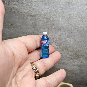 Puede incluir: Una botella de pl&aacute;stico azul en miniatura de refresco Faygo con una tapa blanca. La etiqueta de Faygo es rosa y azul. La botella est&aacute; sostenida en la mano de una persona.