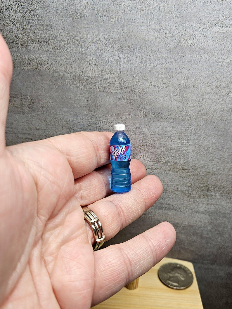 Puede incluir: Una botella de pl&aacute;stico azul en miniatura de refresco Faygo con una tapa blanca. La etiqueta de Faygo es rosa y azul. La botella est&aacute; sostenida en la mano de una persona.
