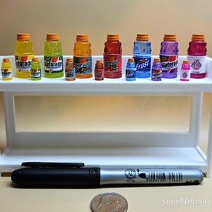 Pu&ograve; includere: Una collezione di bottiglie di Gatorade in miniatura di vari colori, tra cui giallo, verde, arancione, rosso, blu, viola e rosa, esposte su una mensola bianca. Un pennarello nero e una moneta sono in primo piano.