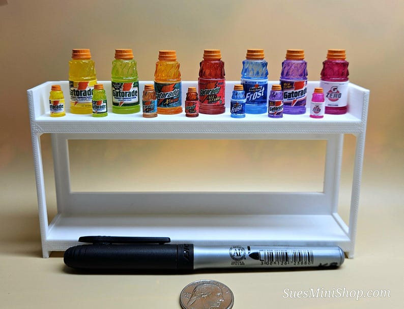 Pu&ograve; includere: Una collezione di bottiglie di Gatorade in miniatura di vari colori, tra cui giallo, verde, arancione, rosso, blu, viola e rosa, esposte su una mensola bianca. Un pennarello nero e una moneta sono in primo piano.