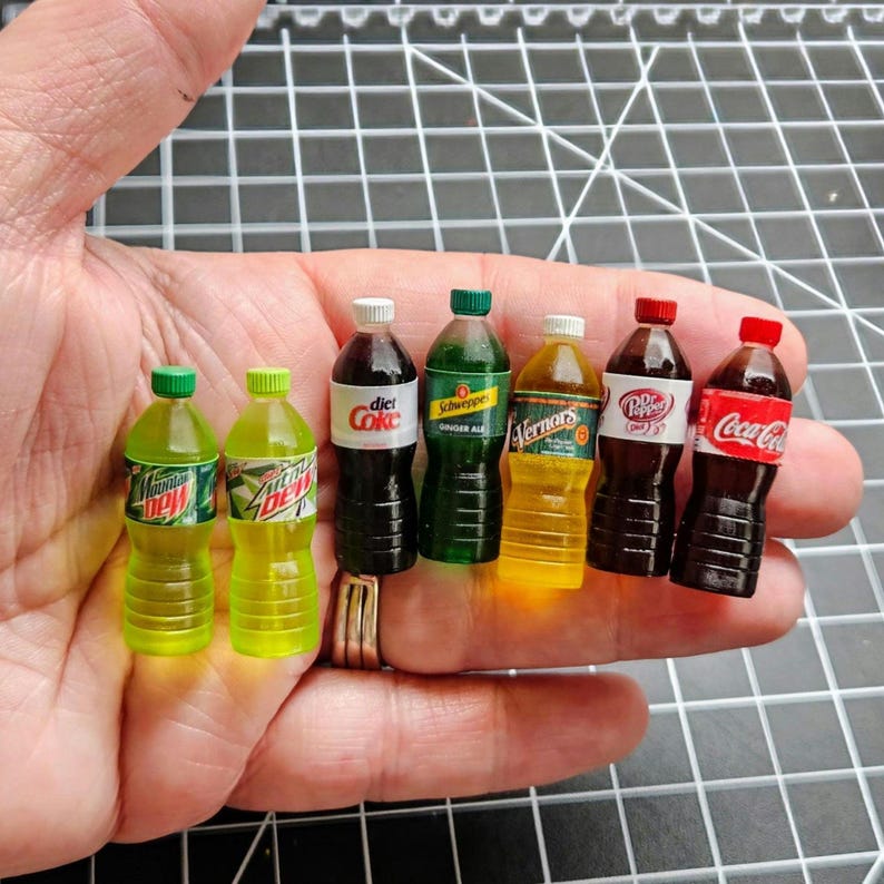 Puede incluir: Se muestran botellas de refrescos en miniatura, incluyendo Mountain Dew, Diet Coke, Schweppes y Coca-Cola. Cada botella tiene una tapa y una etiqueta de diferente color. Las botellas se sostienen en una mano, mostrando la peque&ntilde;a escala de los art&iacute;culos.