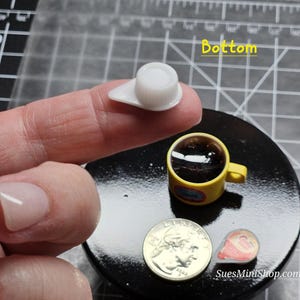 Może przedstawiać: Miniaturowa biała ceramiczna filiżanka z ż&oacute;łtym uchwytem, wypełniona ciemnym płynem, stojąca na czarnej powierzchni. Filiżanka znajduje się obok srebrnej monety i małego czerwono-białego rożka lodowego. Tekst "Bottom" jest napisany na ż&oacute;łto na czarnej powierzchni.