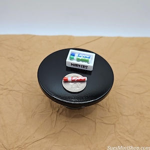 May include: A miniature black table with a white box of markers labeled "Markers" and a red and white Expo marker on a silver coin.