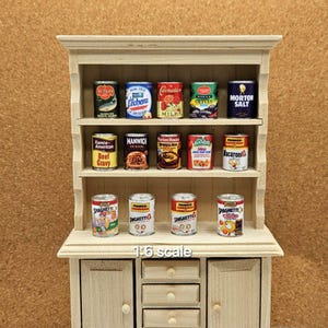 May include: A miniature wooden cabinet with three shelves, filled with small cans of food. The cans feature labels like "Olives," "Spaghetti," and "Beef Gravy." The cabinet is labeled "1:6 scale."