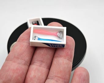 Könnte beinhalten: Eine weiße Miniaturbox mit einer blauen und rosa Rasierklinge darin. Die Box ist mit "Venus" beschriftet und hat einen weißen Hintergrund mit blauer Schrift.