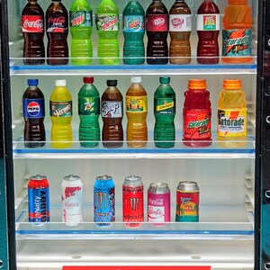 Pu&ograve; includere: Un frigorifero in miniatura pieno di bottiglie e lattine di bevande varie. La selezione comprende Coca-Cola, Pepsi, Gatorade e bevande energetiche. Il testo "1:6 scale Beverages" &egrave; visualizzato in basso.