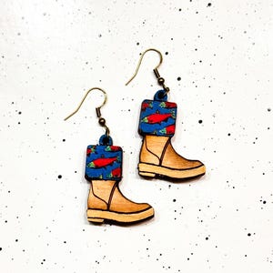 May include: A pair of wooden earrings shaped like boots with a salmon print design. The boots are brown with blue and red accents.