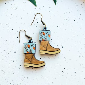 May include: A pair of wooden earrings shaped like boots with a blue and orange patterned design. The boots are brown with a white sole.