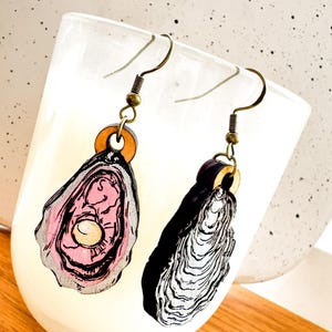 May include: A pair of wooden earrings shaped like oysters. One earring is painted pink with a white pearl inside, while the other is painted black and white.