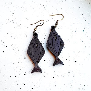 May include: A pair of dangle earrings featuring a dark gray fish design. The fish have a detailed texture and are suspended from gold-toned hooks.