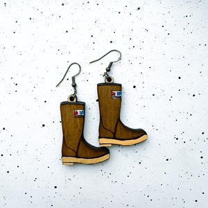 May include: A pair of brown wooden earrings shaped like rain boots. The boots have a white stripe and the text "Sea-Tugs" on the side.