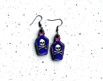 Peut inclure: Une paire de boucles d'oreilles pendantes en forme de flacon de potion. Chaque flacon est coloré dans des tons de violet et de bleu avec un motif de crâne et d'os croisés blancs sur le devant. Les boucles sont fixées à des crochets en métal sombre.