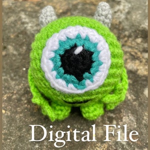 May include: Green crocheted plush toy with a single large eye. The eye is made of teal, white, and black yarn. The toy has two small gray horns on top of its head. The toy is sitting on a gray and brown textured surface. Digital File.