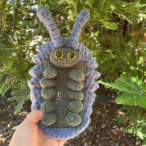 May include: A crocheted pill bug plush toy featuring a light purple outer shell and a dark olive green underside. The creature has two long antennae, a rounded head, and multiple small legs along its body, finished with two large, shiny gold eyes.