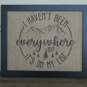May include: A framed burlap print with the text "I haven't been everywhere but it's on my list" in a circle with a mountain range and pine trees.