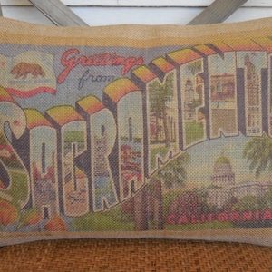 May include: Rectangular burlap pillow with a vintage postcard design. The pillow features the words "Greetings from Sacramento California" in colorful block letters. The design includes images of landmarks and a California flag.