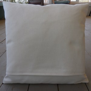 May include: A plain white pillow with a white fabric backing. The pillow is lying flat on a wooden floor.