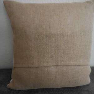 May include: A natural-colored burlap pillow cover with a subtle texture. The pillow cover has a seam down the center and is made of a woven fabric.