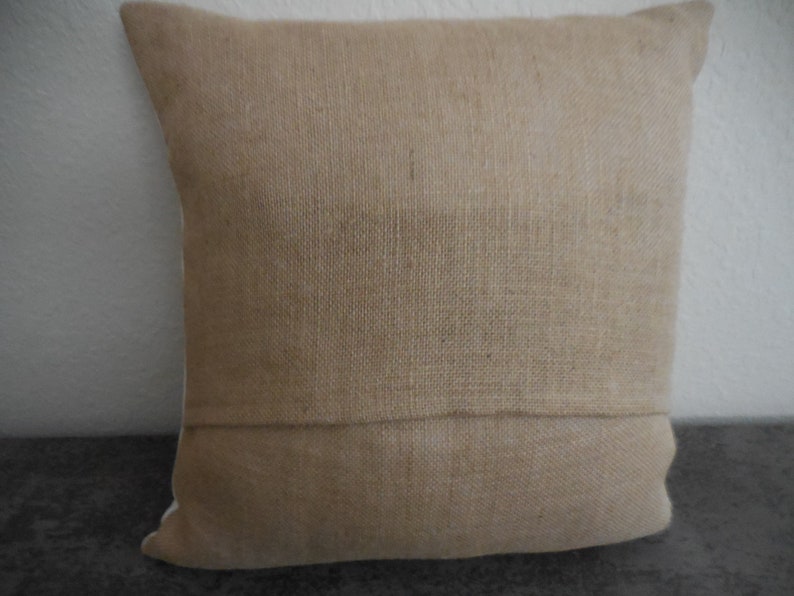 May include: A natural-colored burlap pillow cover with a subtle texture. The pillow cover has a seam down the center and is made of a woven fabric.