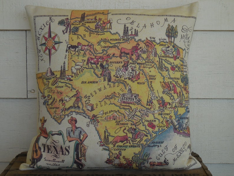 May include: A vintage-style map of Texas on a beige pillow. The map is yellow and features illustrations of various landmarks, animals, and cities. The text "Texas" is written in a large font at the bottom of the map, along with the words "700 miles".