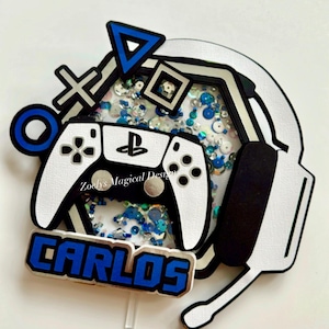 May include: A white and black gaming-themed cake topper with a controller, headset, and name "CARLOS" in blue. The topper includes blue and silver confetti and various gaming symbols.