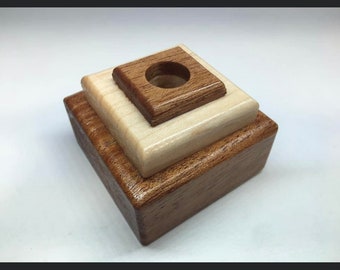 May include: A wooden pen holder made of three stacked squares. The top square is a darker wood with a round hole in the center. The middle square is a lighter wood. The bottom square is a darker wood.