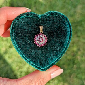May include: A gold pendant featuring a cluster of red and clear gemstones in a floral pattern. The pendant is presented in a heart-shaped, dark green velvet jewellery box. The gemstones are encircled by a halo of smaller, clear stones.
