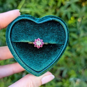 May include: A pink flower-shaped ring with small white diamonds surrounding a larger pink stone, set in a gold band. The ring is displayed in a green velvet heart-shaped ring box.