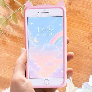Pode incluir: Uma capa de telefone rosa com orelhas de gato envolve um smartphone exibindo um papel de parede pastel com arco-íris e nuvens. A tela mostra a hora 12:37 na quinta-feira, 20 de abril. A capa tem um botão home e a frase "Press home to unlock".