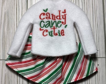 May include: A white elf-sized long-sleeved shirt with the text "Candy Cane Cutie" embroidered in red and green. The shirt is paired with a red and green striped skirt.