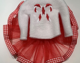 May include: A white felt outfit featuring a red candy cane design on the front. The outfit includes a red tulle skirt with a gingham ribbon trim. The festive ensemble is designed for a doll or small figure.
