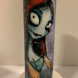 May include: A stainless steel tumbler with a colorful illustration of a character with red hair, blue eyes, and a stitched-up face. The background is a purple and white splatter pattern.