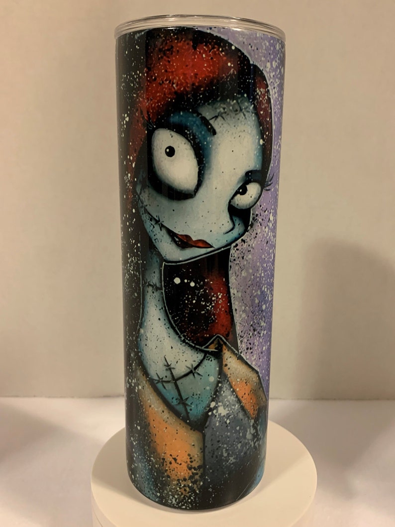 May include: A stainless steel tumbler with a colorful illustration of a character with red hair, blue eyes, and a stitched-up face. The background is a purple and white splatter pattern.