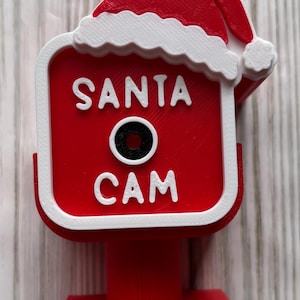 May include: A red and white 3D printed Santa Cam ornament with a square frame, a Santa hat topper, and a black circular lens in the center. The text SANTA CAM is embossed in white on the red face of the square device, which is mounted on a red base.