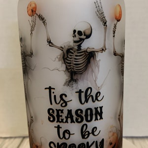 May include: White frosted glass jar with a bamboo lid. The jar has a Halloween-themed design with two skeletons holding pumpkins and the text "Tis the season to be spooky".