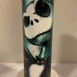May include: A black and white stainless steel tumbler with a graphic of Jack Skellington from the Nightmare Before Christmas movie. The tumbler has a teal and black background with a white and black image of Jack Skellington. The image also includes a black cat with yellow eyes.
