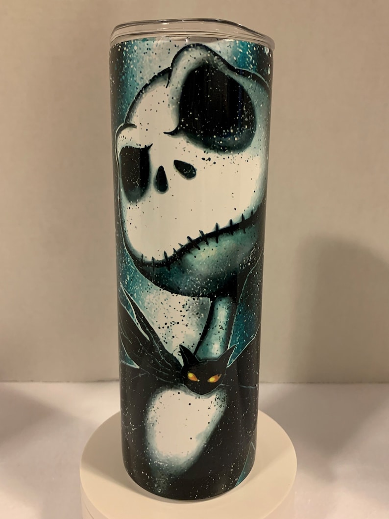 May include: A black and white stainless steel tumbler with a graphic of Jack Skellington from the Nightmare Before Christmas movie. The tumbler has a teal and black background with a white and black image of Jack Skellington. The image also includes a black cat with yellow eyes.