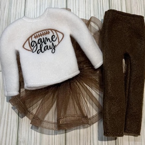 May include: A white elf-sized shirt with a brown football and the text "game day" embroidered on the front. The shirt is paired with a brown tulle skirt and brown fleece pants.