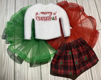 May include: A white elf-sized sweater with the text "merry christmas" embroidered in red. The sweater is paired with a red tulle skirt and a black and red plaid skirt with a green Christmas tree pattern.