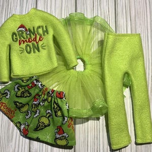 May include: A green Grinch-themed outfit for a doll, including a long-sleeved shirt with the text "Grinch mode on", a green tulle skirt, green pants, and a green skirt with a Grinch print.