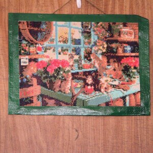 May include: A green framed cross-stitch picture depicting a colorful floral arrangement in a window box with a view of a garden scene.