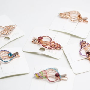 May include: Several seashell-shaped hair clips with rose gold metal frames and colorful rhinestone accents. Each clip is attached to a white card. The clips feature various colors, including pink, blue, and gold.