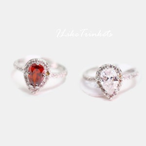 May include: Two pear-shaped engagement rings with a halo setting. The ring on the left has a red gemstone, while the ring on the right has a clear gemstone.