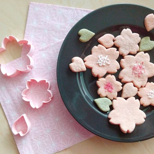 Op de afbeelding: Een bord met roze en groene suikerkoekjes in de vorm van kersenbloesems. De koekjes zijn versierd met witte sprinkles.