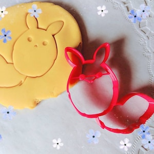 Puede incluir: Un cortador de galletas de plástico rojo con forma de conejo. El cortador está sobre una superficie blanca con un recorte de masa de galleta amarilla en el fondo.