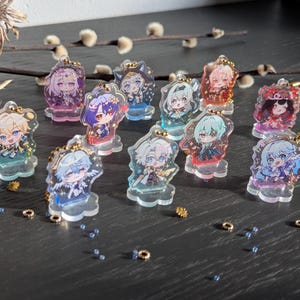 Könnte beinhalten: Eine Sammlung von zwölf kleinen, durchsichtigen Acryl-Aufstellern mit farbenfrohen Illustrationen im Anime-Stil. Jede Figur steht auf einem wolkenförmigen Sockel und hat oben eine kleine goldfarbene Metallöse zum Aufhängen als Anhänger.
